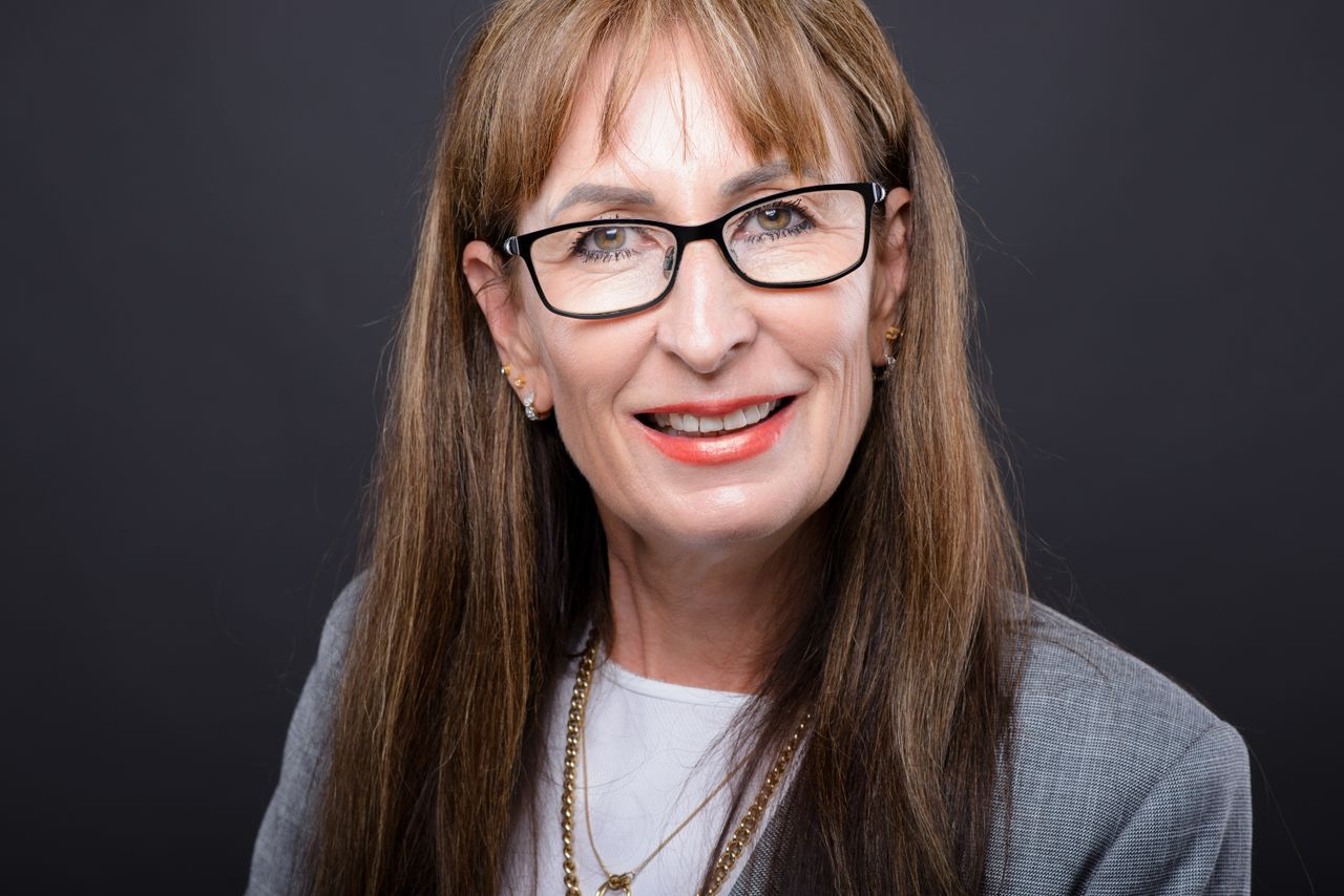 A senior woman smiling in eyeglasses and a gray coat, with a confident expression.